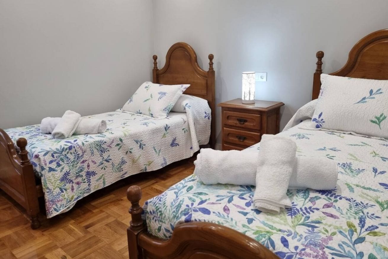 A room with two wooden beds, each with a floral bedspread and matching pillows, a nightstand with a lamp, and a wooden floor.