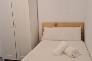 A single bed with a wooden headboard and white bedding, a white wardrobe, and a rolled towel on the bed.