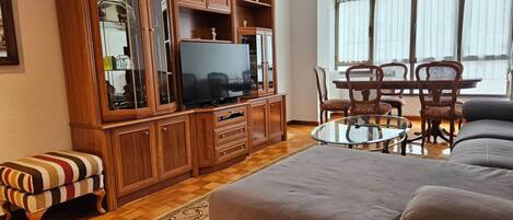 A living room with a television, wooden cabinet, striped ottoman, and a glass coffee table.