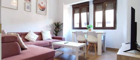 Ein Wohnzimmer mit einem rosafarbenen Sofa, einem weißen Couchtisch und einem Fernseher auf einem Holzständer. Zwei Fenster mit braunen Rahmen und eine Pflanze auf der Fensterbank sind ebenfalls zu sehen.