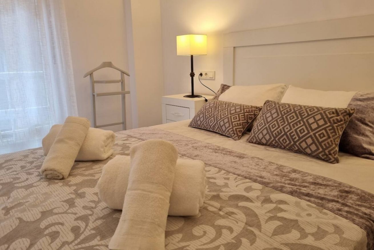 A neatly made bed with patterned pillows, a bedside lamp, and rolled towels on a patterned bedspread.