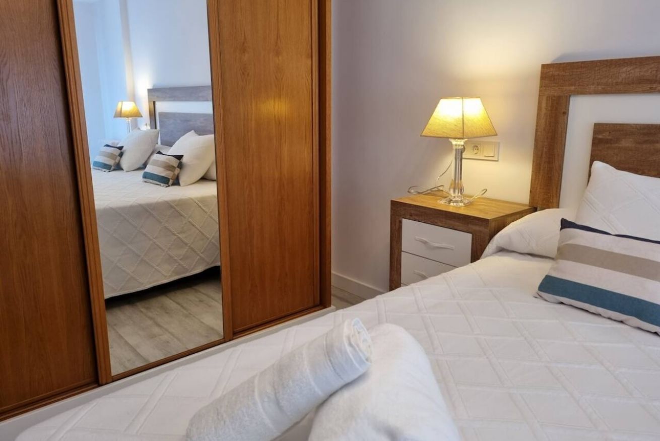 Habitación de hotel con cama, mesita de noche con lámpara, armario de madera y un espejo que refleja la habitación.
