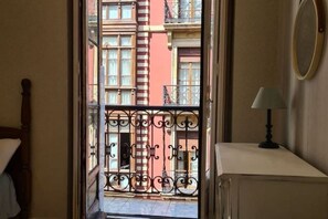 A narrow hallway leading to a balcony with wrought iron railings, a bed, a nightstand, and a lamp.