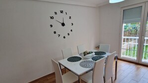 A dining area with a white table, four chairs, a wall clock, and a window with a view of greenery.