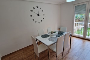 A dining area with a white table, four chairs, a wall clock, and a window with a view of greenery.