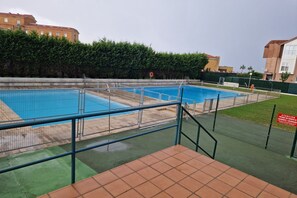 Una piscina con una valla de seguridad, una zona empaveda al frente y edificios al fondo.