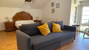 A bedroom with a bed, a grey sofa with yellow cushions, wooden nightstands, and a window with a view.