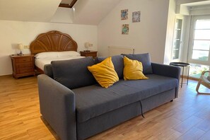 Une chambre à coucher avec un lit, un canapé gris agrémenté de coussins jaunes, des tables de chevet en bois et une fenêtre donnant sur un paysage agréable.