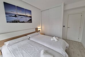Un lit double avec du linge de lit blanc, une table de chevet avec une lampe et une œuvre d’art fixée au mur représentant un paysage marin.