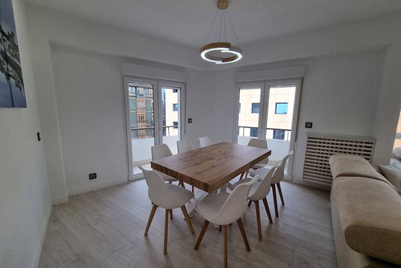 A modern dining area with a wooden table and white chairs, large windows, and a beige sofa.