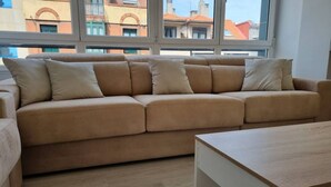 Un canapé beige agrémenté de plusieurs coussins, dans une pièce dotée de grandes fenêtres et d’un plancher en bois.