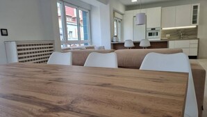 Une chambre d’hôtel moderne avec une table à manger en bois, des chaises blanches, un canapé, un coin cuisine et une grande fenêtre.