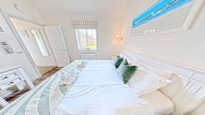 A hotel room with a bed, a window with curtains, a wall-mounted lamp, and a painting. - Stunning Beach House - Sleeping 4 in 2 Bedrooms - Pool and Beach Access Included. (Primrose Valley)