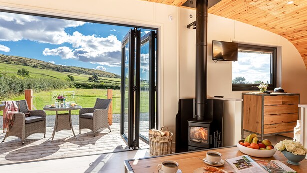 A cozy room with a wooden ceiling, a large window offering a scenic view of green hills, a fireplace, wicker furniture, and a dining table set with breakfast. - Little Heaning (Grange-over-Sands)