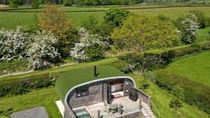 A modern, curved wooden structure with a glass enclosure, a patio with seating, and a view of a lush green landscape. - Little Heaning (Grange-over-Sands)