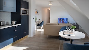 A modern kitchen with dark cabinets, a microwave, and a toaster. There is a dining area with a round table, blue chairs, and a TV in the background. - Maison Douaud Vannes (Vannes)