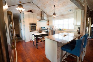 Une cuisine spacieuse avec un plancher en bois, des armoires blanches, un îlot central doté de tabourets et un éclairage suspendu.