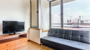 Un salón moderno con un televisor de pantalla plana sobre un mueble de madera, un sofá gris y grandes ventanales que ofrecen vistas de la ciudad.