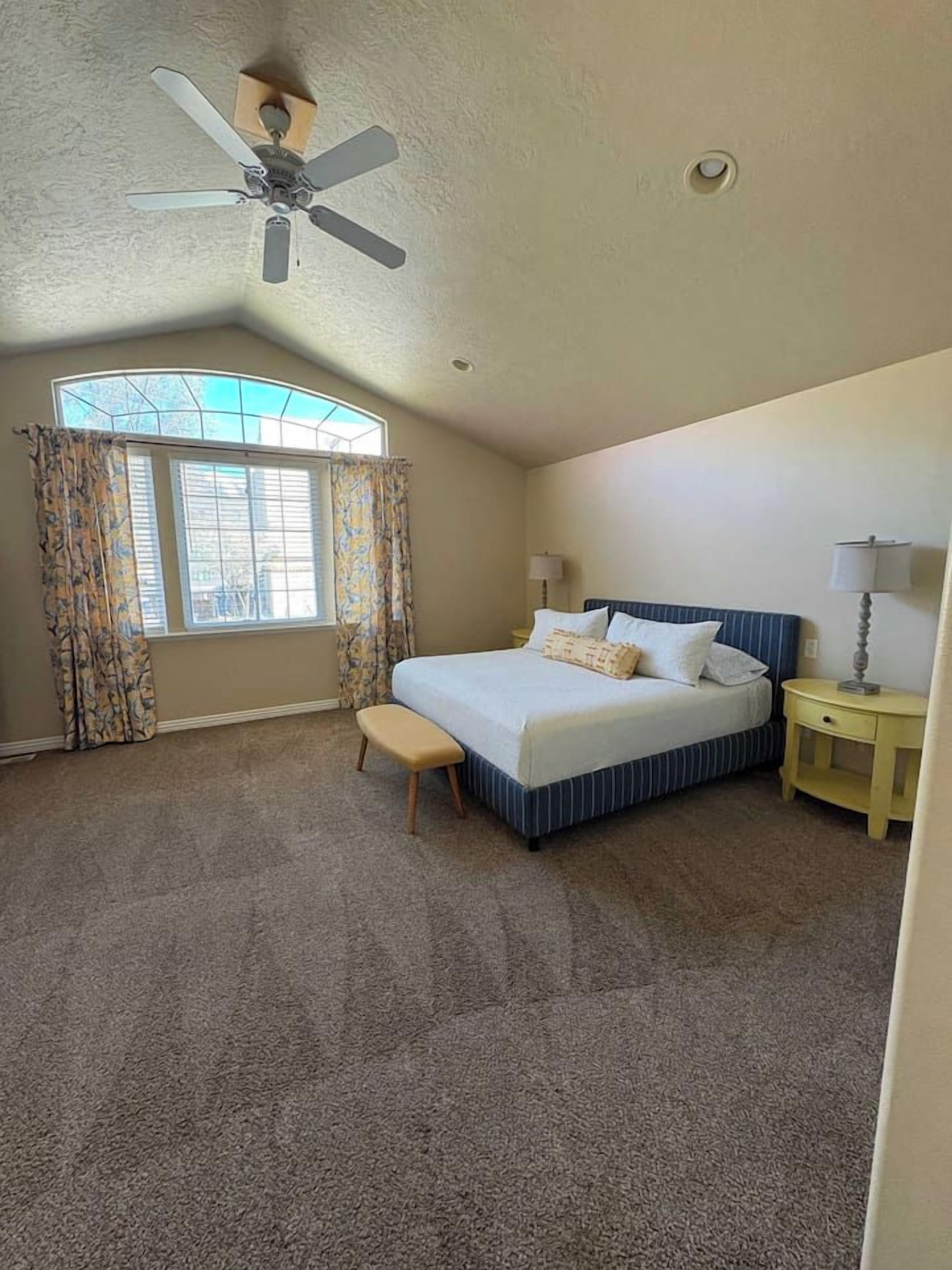 A bedroom with a bed, a bench, a nightstand, a ceiling fan, and a window with curtains.