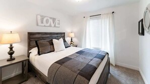 Un dormitorio con cama, dos mesitas de noche con lámparas, un espejo en la pared y una ventana con cortinas.