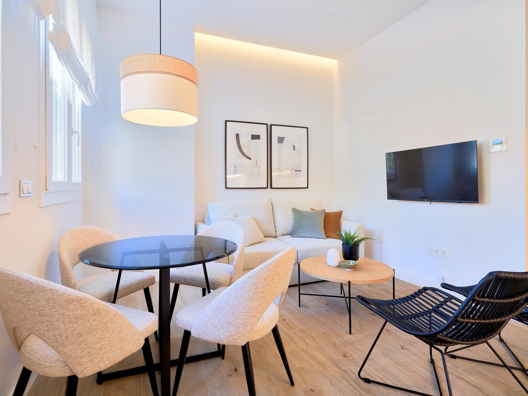 Uma sala de estar moderna com uma mesa de vidro redonda, duas poltronas brancas, uma cadeira de vime preta, uma pequena mesa redonda com uma planta, um sofá com almofadas e uma TV de tela plana na parede.