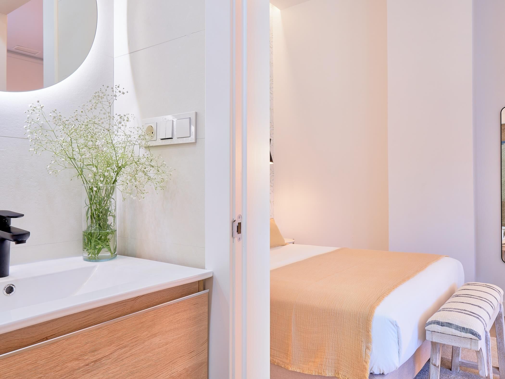 Un dormitorio moderno con un tocador de madera, un lavabo blanco, un espejo, una mesita de noche y una cama con una manta color beige, junto a un taburete pequeño.