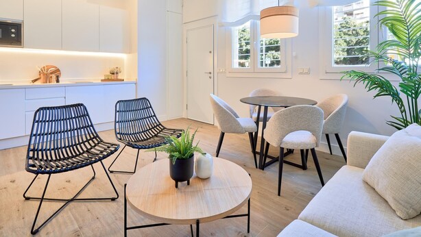Ein modernes Wohnzimmer mit einem runden Holztisch, zwei schwarzen Gestellstühlen, einem beigen Sofa und einer großen Topfpflanze.