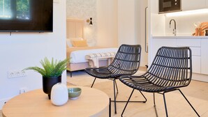 Ein modernes Hotelzimmer mit einem Bett, einem Fernseher, einer Essecke mit zwei Stühlen, einem kleinen Tisch, einer Pflanze und einer Küchenzeile inklusive Mikrowelle.