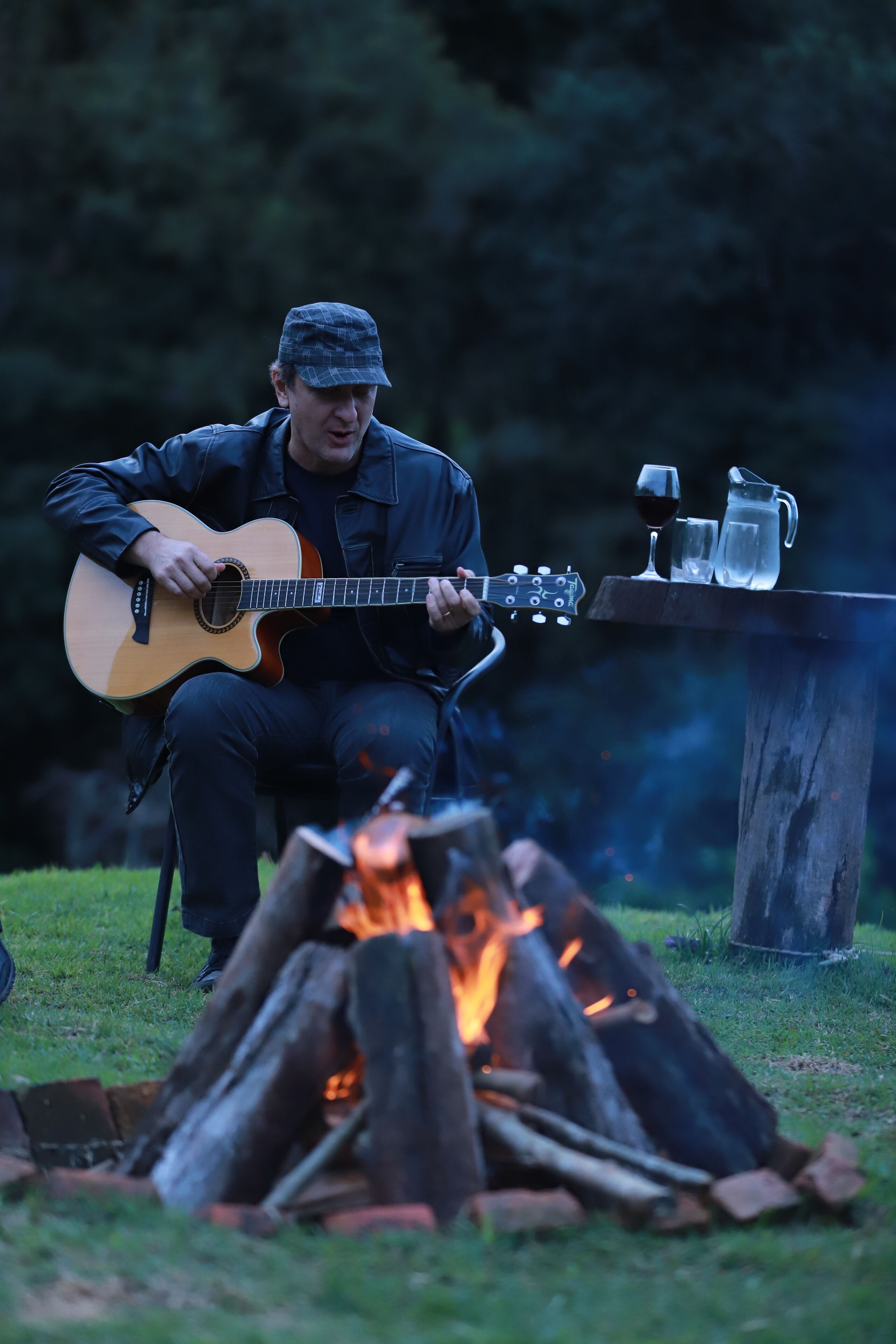 Une personne joue de la guitare acoustique près d’un feu de camp. Une table se trouve à côté d’elle, sur laquelle sont posés un verre de vin rouge et une carafe.
