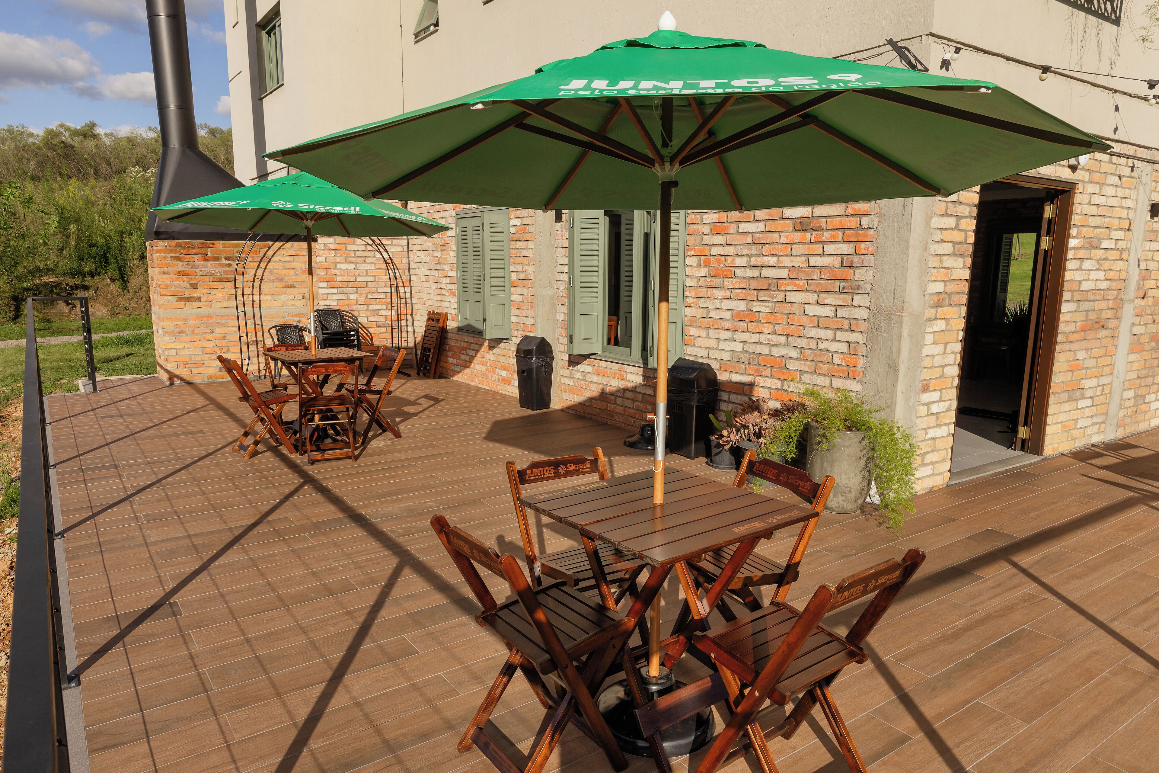 Eine Terrasse im Freien mit Holztischen und -stühlen, grünen Sonnenschirmen und einer Backsteinmauer.