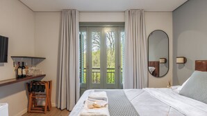 A hotel room with a bed, a wooden chair, a shelf with bottles and glasses, a mirror, and a view of greenery through the door. - Pousada Calza (Bento Goncalves)