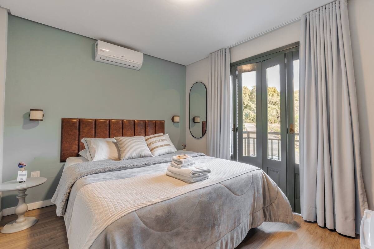 Un dormitorio con cama, mesitas de noche, aire acondicionado, ventana con cortinas y vistas al exterior.