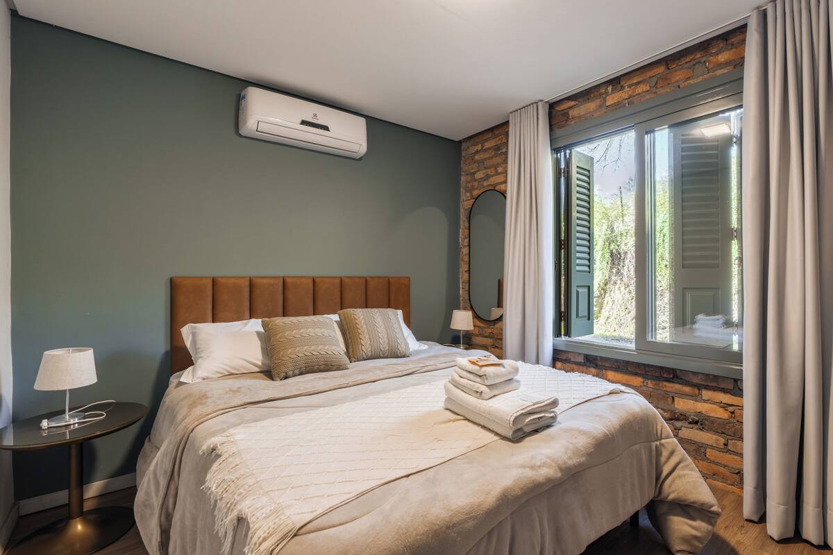 Una cama bien tendida con cabecera, una lámpara de noche y una ventana con cortinas.