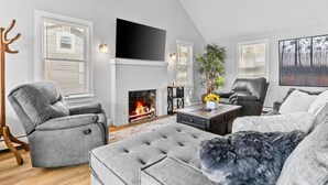 A modern living room with a fireplace, grey sofas, a flat-screen TV, and a large window with blinds. - Beach House · Beautiful Beachfront House, Close to Burlington! (Colchester)