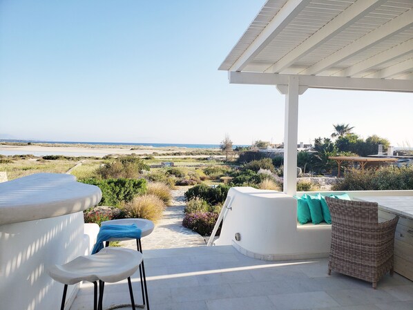 Une terrasse dotée de mobilier blanc, offrant une vue sur la plage, et un espace abrité avec un fauteuil en osier.