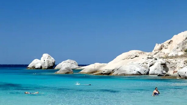 Une plage avec une eau turquoise et limpide, de gros rochers blancs et quelques personnes qui se baignent.
