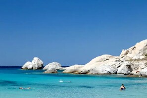 Une plage avec une eau turquoise et limpide, de gros rochers blancs et quelques personnes qui se baignent.