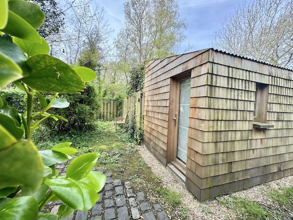 A small wooden structure with a door and a window, surrounded by greenery and a cobblestone path.