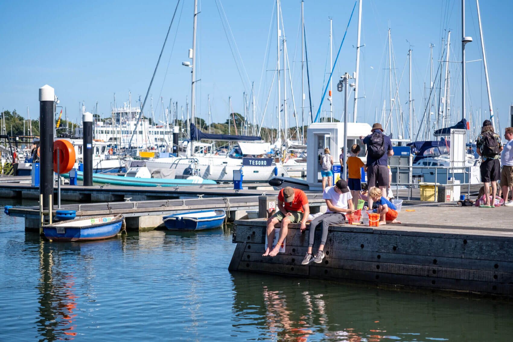 A marina with boats docked, people sitting on a dock, and a clear sky.