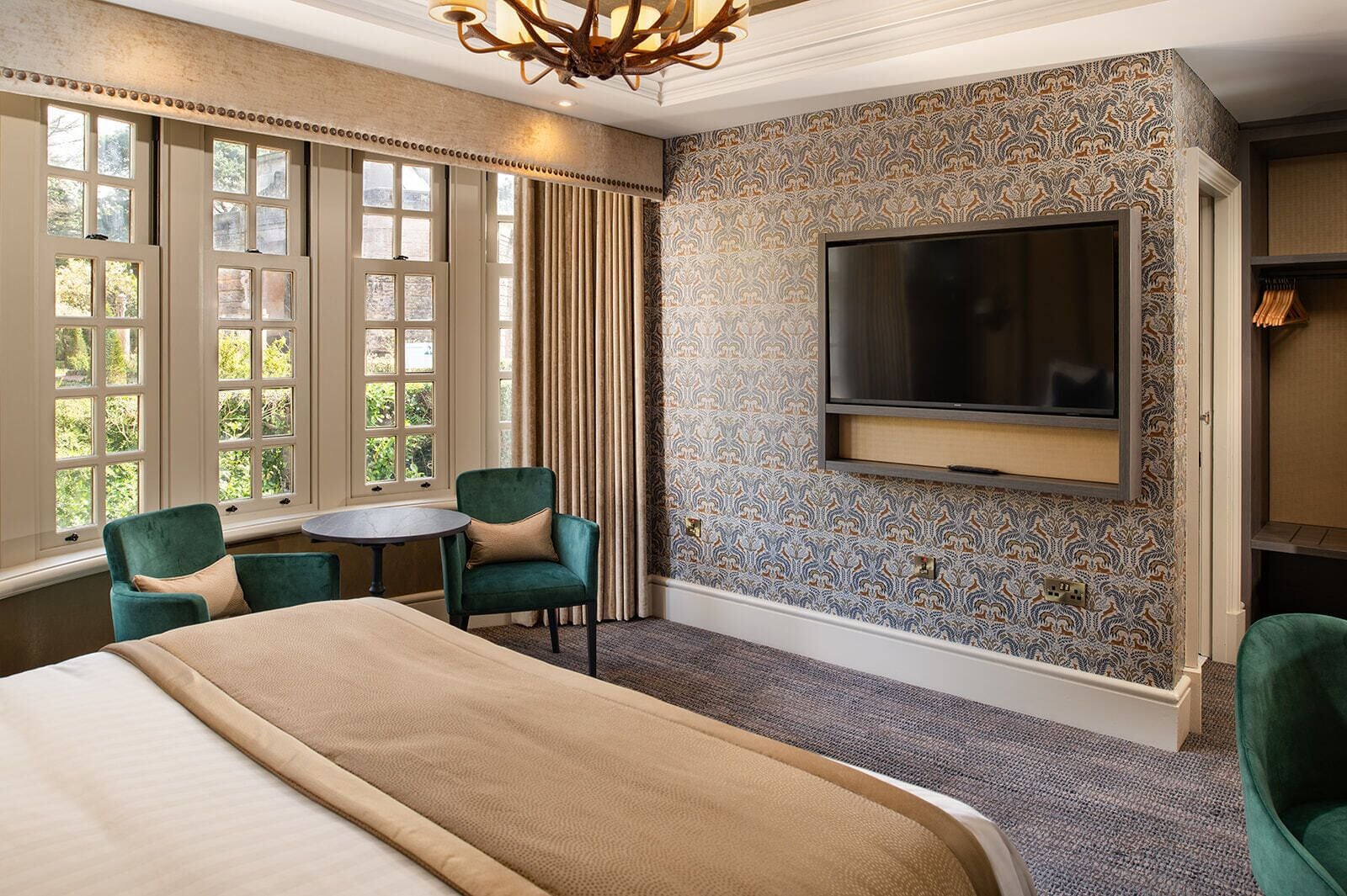 Una habitación de hotel con una cama grande, dos sillones verdes, una mesa redonda, un televisor montado en la pared y un papel tapiz estampado.