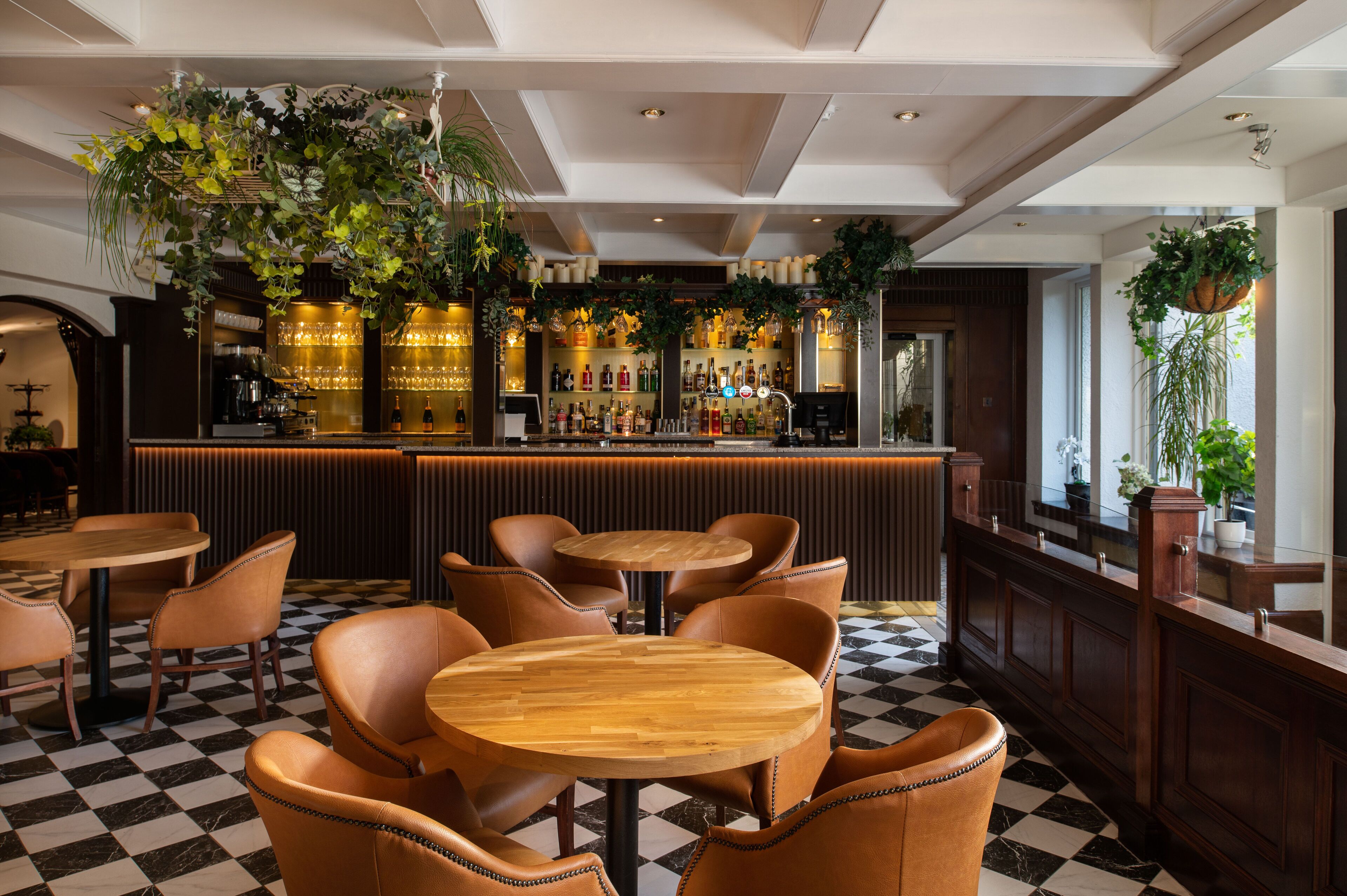 Una zona de bar con mesas y sillas de madera, suelo a cuadros y plantas colgantes.