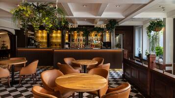Una zona de bar con mesas y sillas de madera, suelo a cuadros y plantas colgantes.