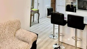 Ein modernes Wohnzimmer mit einem gemusterten Sofa, einem Essbereich mit Barhocker und einer Küche mit weißen Geräten.