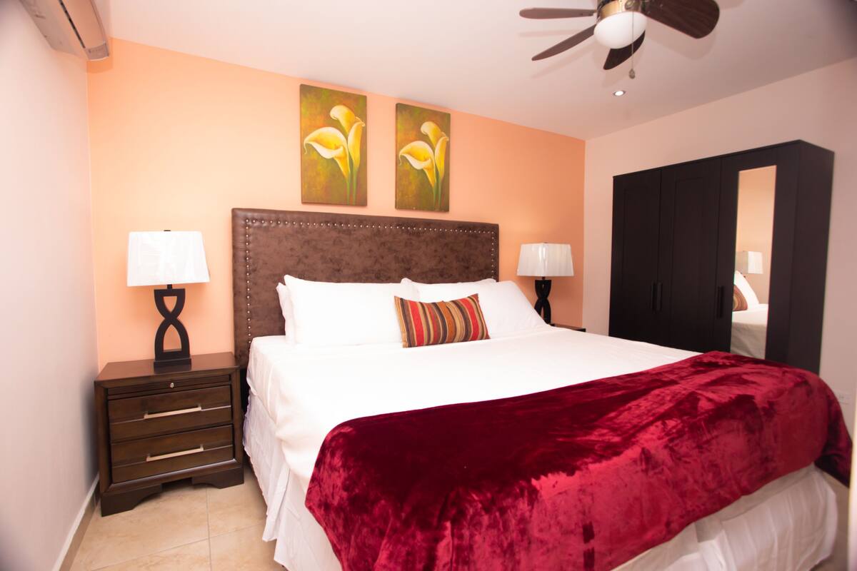 A bedroom with a large bed, two wall-mounted paintings, a ceiling fan, a wardrobe, and a nightstand with a lamp.