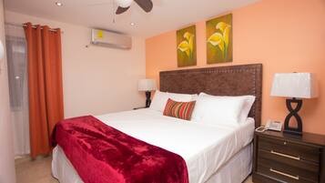 Habitación de hotel con una cama grande, dos aires acondicionados instalados en la pared, un ventilador de techo, una mesita de noche con lámpara y dos cuadros en la pared.