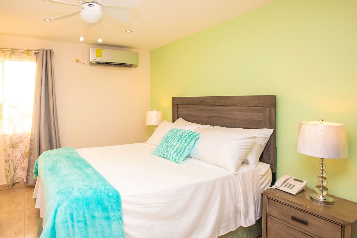 A bedroom with a bed, bedside lamps, a nightstand with a phone, and an air conditioning unit on the wall.