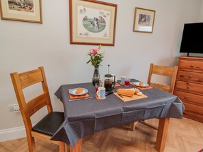 Une table à manger recouverte d’une nappe à pois, deux chaises en bois, un vase avec des fleurs, une cafetière, une théière, des tasses et une assiette avec un croissant.