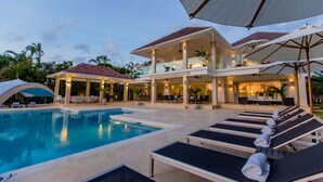 Un espace aménagé au bord de la piscine, avec des chaises longues et des parasols, une grande piscine et un bâtiment à deux étages doté d’un espace extérieur pour s’asseoir et d’une terrasse couverte.