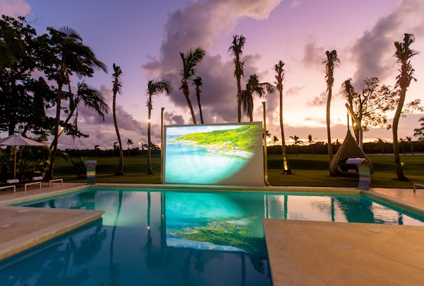 A swimming pool with a large screen displaying a beach scene, surrounded by palm trees and a grassy area.