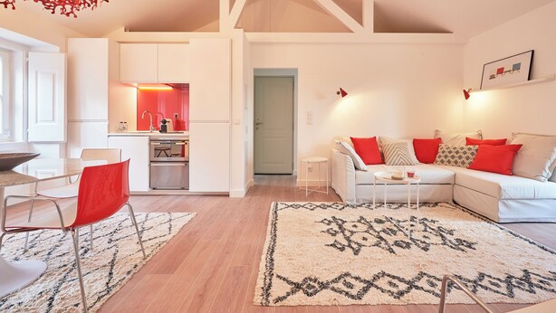 Una sala de estar moderna con un sofá blanco, cojines rojos, un comedor con una silla roja y una cocina al fondo.